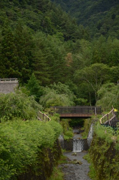 Lush greenery surrounds a small stream flowing beneath a bridge in a mountainous landscape.